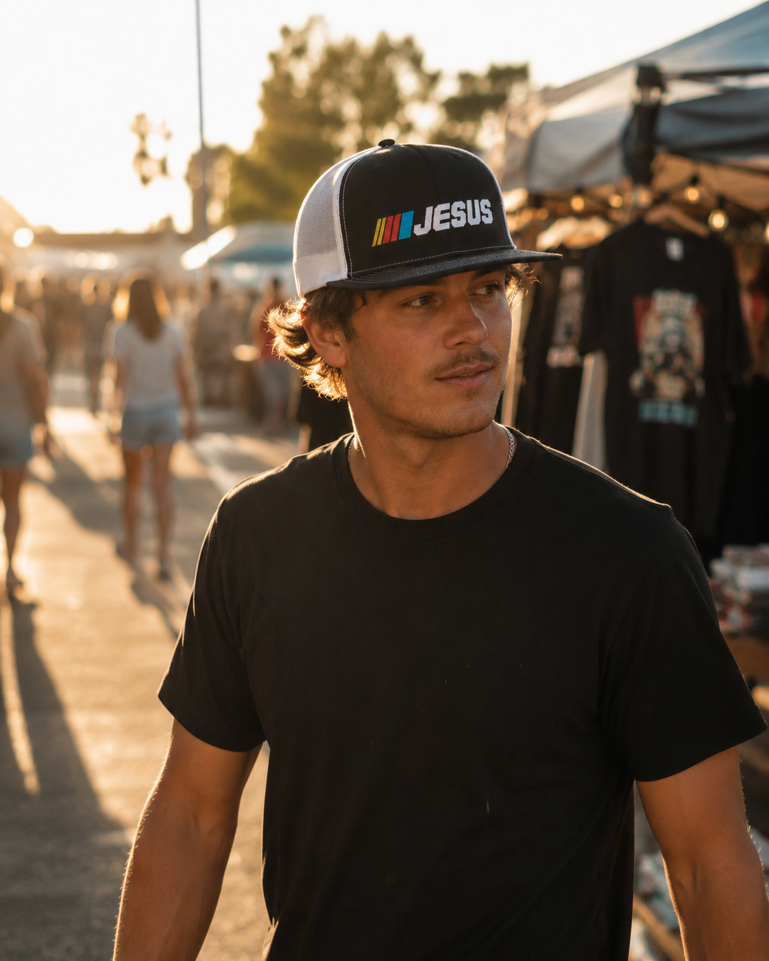 A man wearing a black and white trucker hat with 'M JESUS' printed on the front, standing in an outdoor market setting during golden hour.