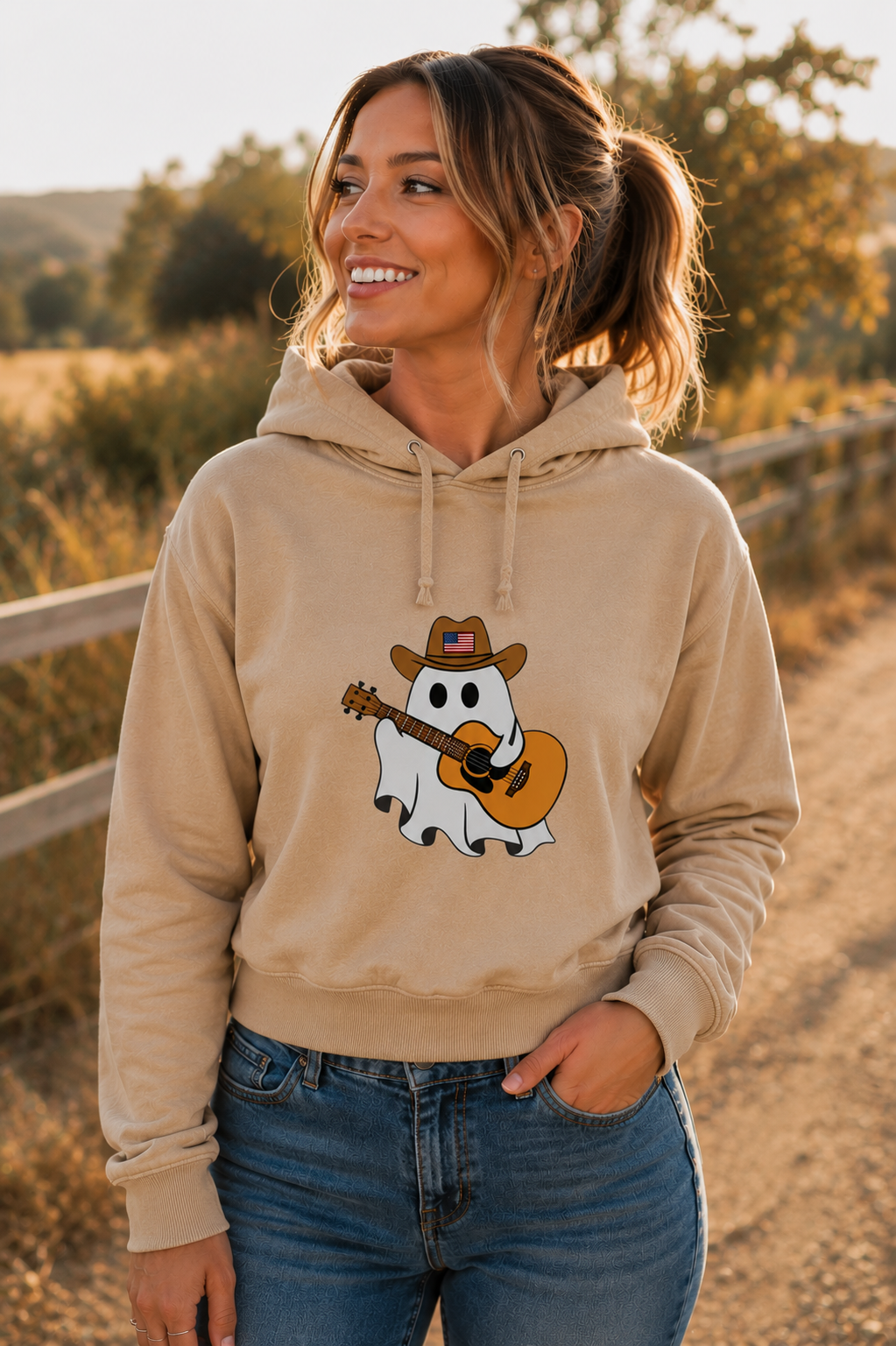 A woman wearing a beige hoodie with a cartoon ghost playing guitar wearing a cowboy hat, paired with blue jeans, standing on a dirt path outdoors.