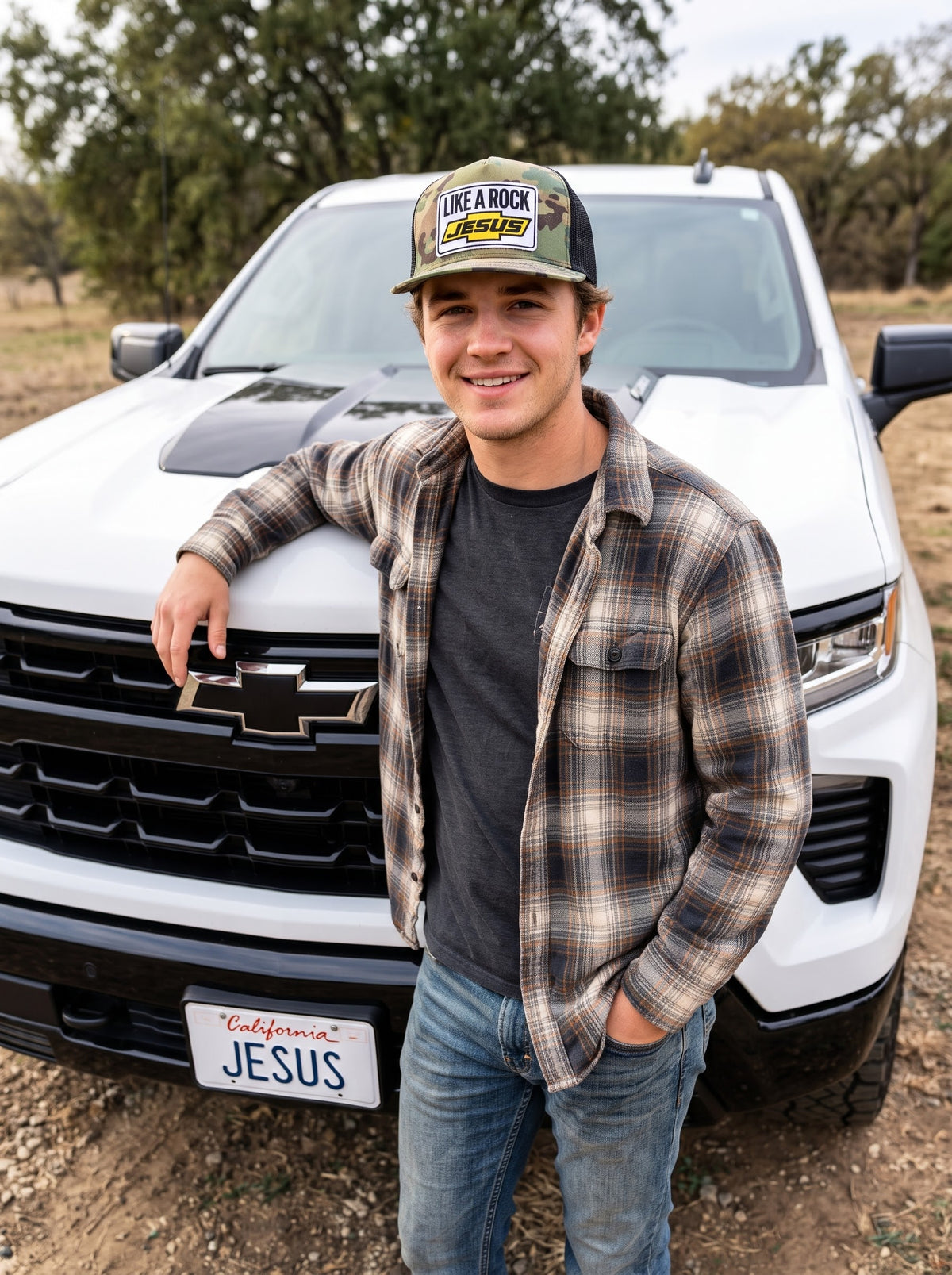 “Man wearing a Like a Rock Jesus camo trucker hat and plaid shirt, leaning against a white pickup truck.”