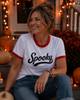 A woman wearing a white ringer T-shirt with red trim and the word 'Spooky' printed in black script, seated on a porch decorated with pumpkins, string lights, and fall foliage.