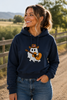 A woman wearing a navy blue hoodie with a cartoon ghost graphic wearing a cowboy hat and playing an acoustic guitar, standing outdoors on a dirt path with a wooden fence and trees in the background.