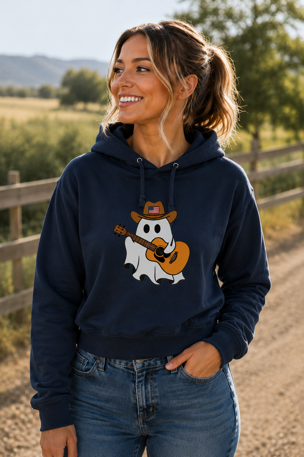 A woman wearing a navy blue hoodie with a cartoon ghost graphic wearing a cowboy hat and playing an acoustic guitar, standing outdoors on a dirt path with a wooden fence and trees in the background.