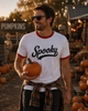 A man wearing a white ringer t-shirt with red trim and a black 'Spooky' graphic, holding a small pumpkin in an outdoor pumpkin patch at sunset.