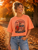 A woman wearing a coral orange graphic t-shirt with a Halloween design that reads 'No Diggity Bout To Bag It Up', paired with distressed blue jeans, standing outdoors on a path covered in fallen autumn leaves.