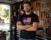 A man standing in a garage workshop wearing a black t-shirt with a graphic of the American flag and the text 'This is a flag. I like it.