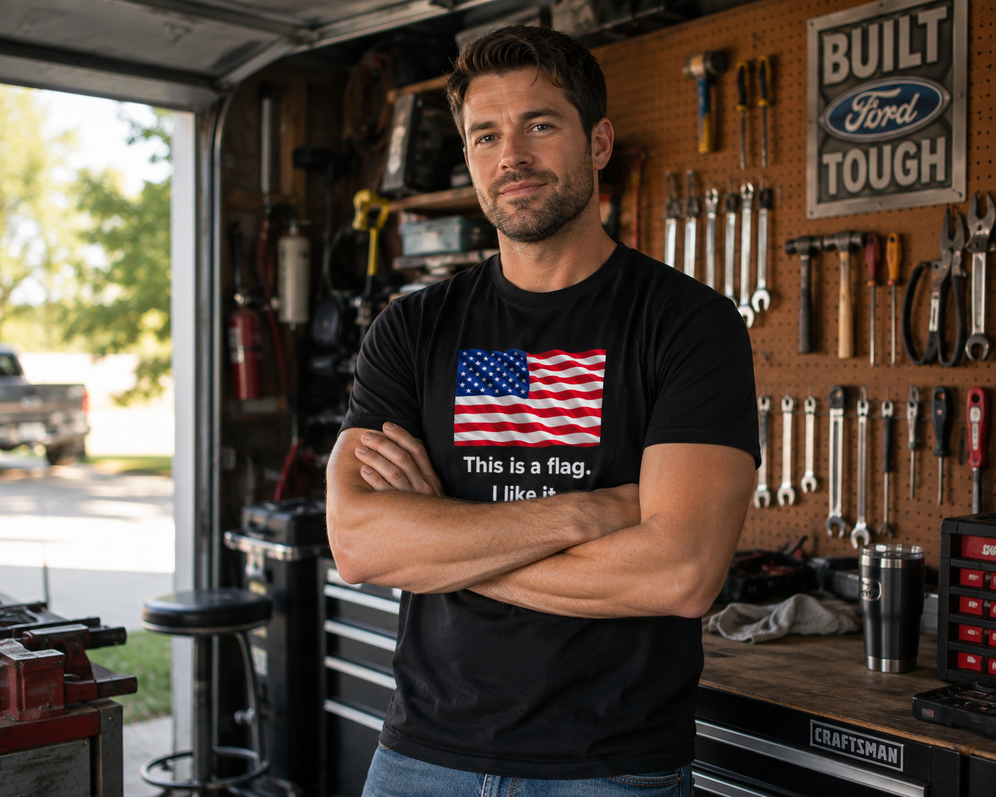 A man standing in a garage workshop wearing a black t-shirt with a graphic of the American flag and the text 'This is a flag. I like it.