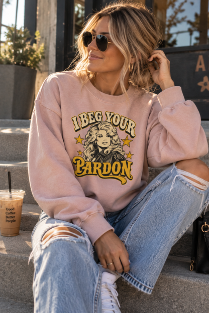 A woman wearing a light pink crewneck sweatshirt with a yellow and black graphic that reads 'LBEG YOUR BARDON' sits on outdoor stone steps. She is wearing ripped light-wash blue jeans, white sneakers, and black sunglasses, with a coffee cup nearby.