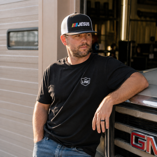 A man wearing a black t-shirt with a white 'The Line' shield logo on the chest and a black-and-white baseball cap with 'JESUS' and a colorful stripe logo, leaning against a vehicle in front of a garage door.