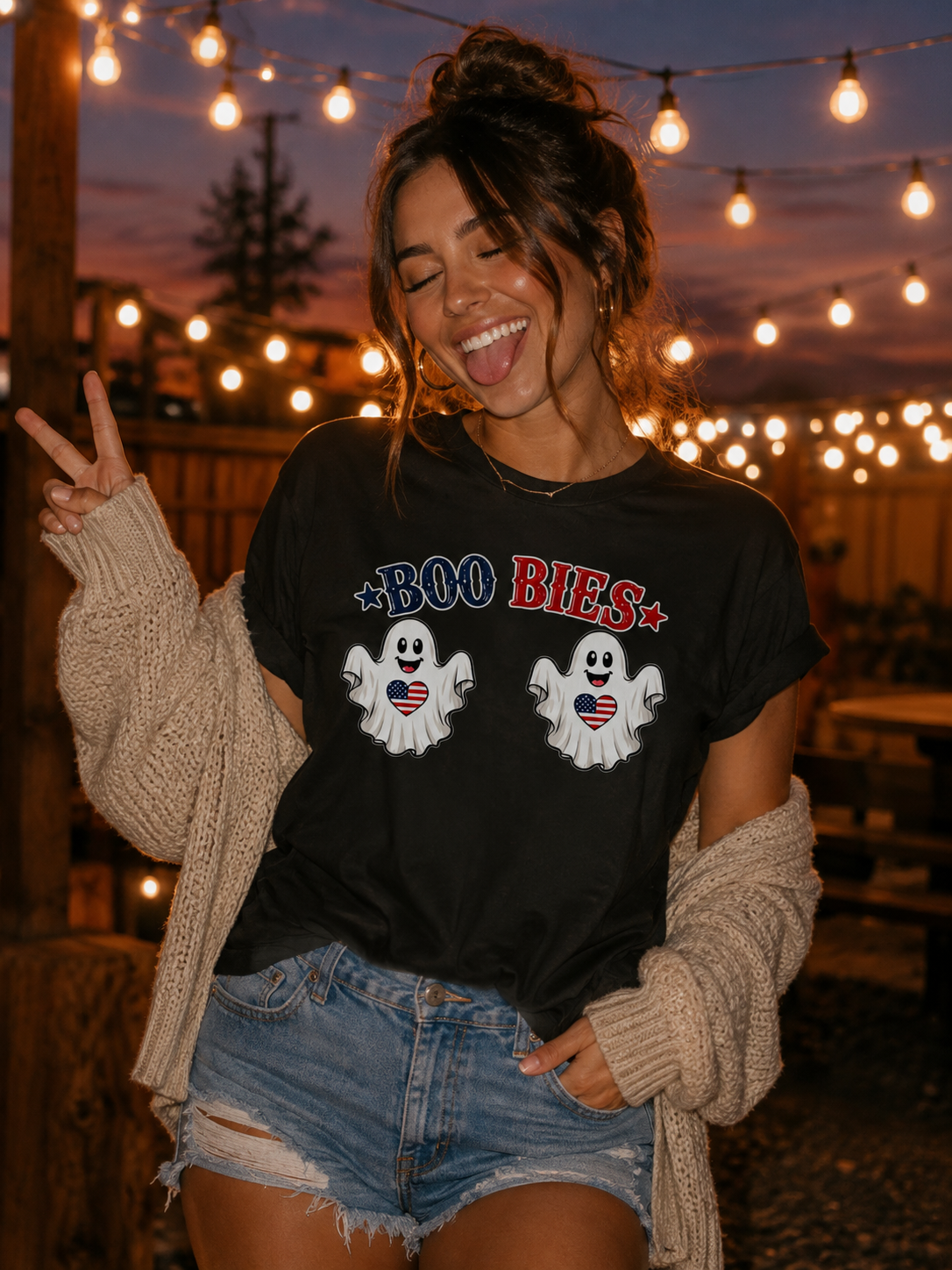 A woman outdoors at dusk wearing a black graphic T-shirt that says 'BOO BIES' with two white ghosts, paired with denim shorts and a beige knit cardigan. Warm string lights are visible in the background.