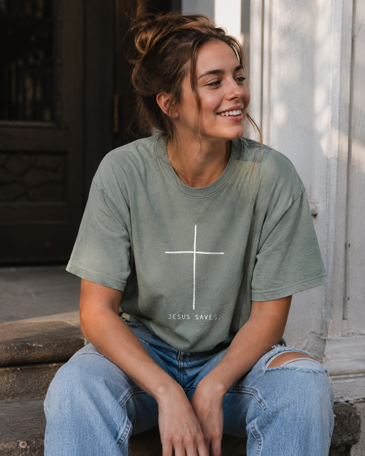 A woman sitting on outdoor stone steps wearing a muted sage green graphic t-shirt with a white cross and the text 'JESUS SAVES' printed on the front, paired with distressed blue jeans.