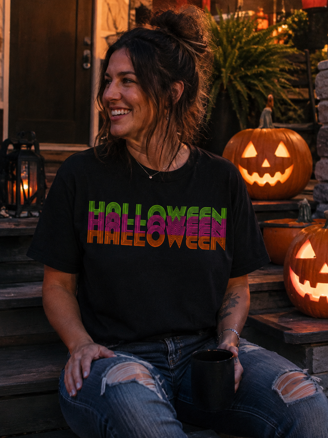 A woman sitting on outdoor wooden steps at night wearing a black graphic t-shirt with 'HALLOWEEN' printed across the chest in a colorful gradient. She is wearing ripped blue jeans and holding a black mug, with glowing jack-o'-lanterns and lanterns in the background.