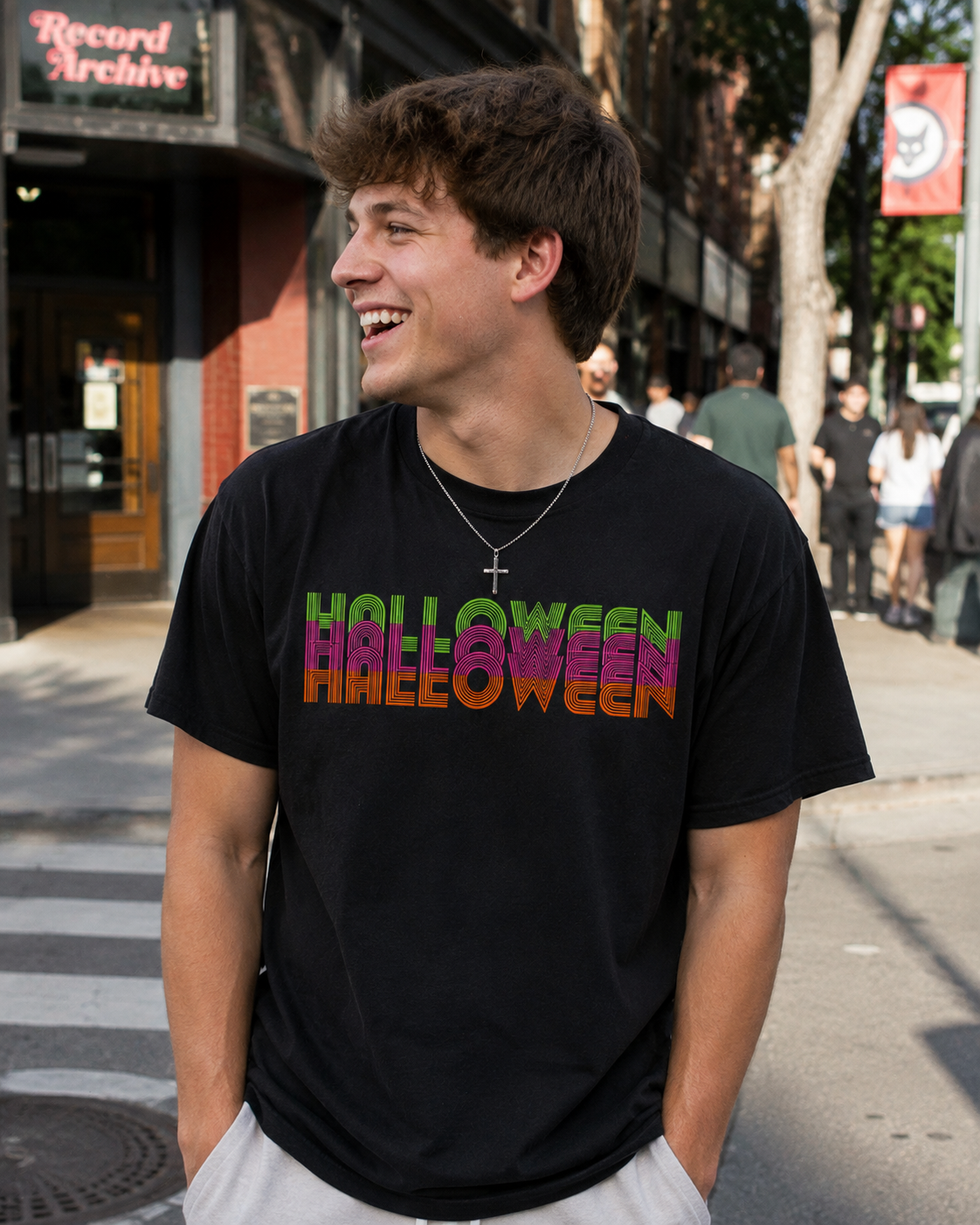 A young man smiling and looking to the side while wearing a black t-shirt with 'HALLOWEEN' printed across the chest in a green-to-purple-to-orange gradient. The background shows a blurred city street scene with a 'Record Archive' storefront.