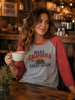A woman in a rustic cafe setting wearing a heather gray and red raglan t-shirt with a 'Make California Great Again' graphic, holding a coffee mug.