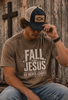 A man wearing a brown graphic t-shirt with the text 'FALL FOR JESUS HE NEVER LEAVES' and a navy blue trucker hat with a fish logo patch, sitting in front of a rustic wooden wall.
