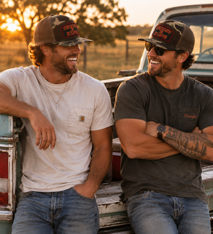 Two men laughing and sitting in the bed of a pickup truck at sunset. Both are wearing camouflage trucker hats with red 'Crazy Dog' logos, sunglasses, and jeans.