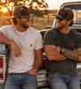 Two men laughing and sitting in the bed of a pickup truck at sunset. Both are wearing camouflage trucker hats with red 'Crazy Dog' logos, sunglasses, and jeans.