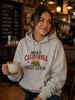 A woman in a cozy cafe setting wearing a light gray heather hoodie with a 'MAKE CALIFORNIA GREAT AGAIN' graphic print in red and blue, holding a ceramic mug.