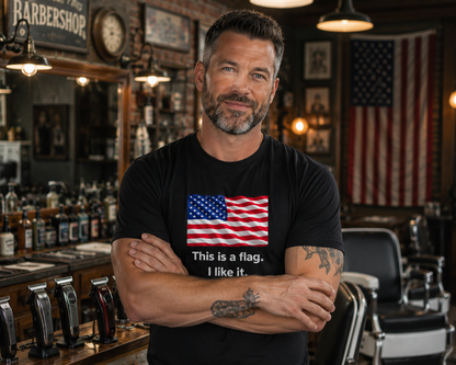 A man with a beard and tattoos stands in a barbershop wearing a black t-shirt with a graphic of the American flag and the text 'This is a flag. I like it.