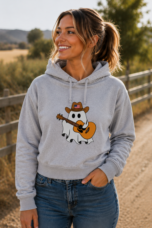 A woman wearing a light gray hoodie with a graphic of a ghost playing an acoustic guitar while wearing a cowboy hat, set against a blurred outdoor background of a dirt path and fence.