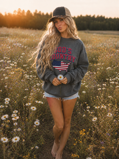 A woman standing in a sunlit field of wildflowers, wearing a dark gray sweatshirt with 'God's Country' and an American flag graphic, denim shorts, and a trucker hat.