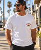 A man wearing a white crewneck pocket t-shirt with a California grizzly bear logo on the chest, paired with a camouflage cap and sunglasses, standing on a sunny beachside boardwalk.