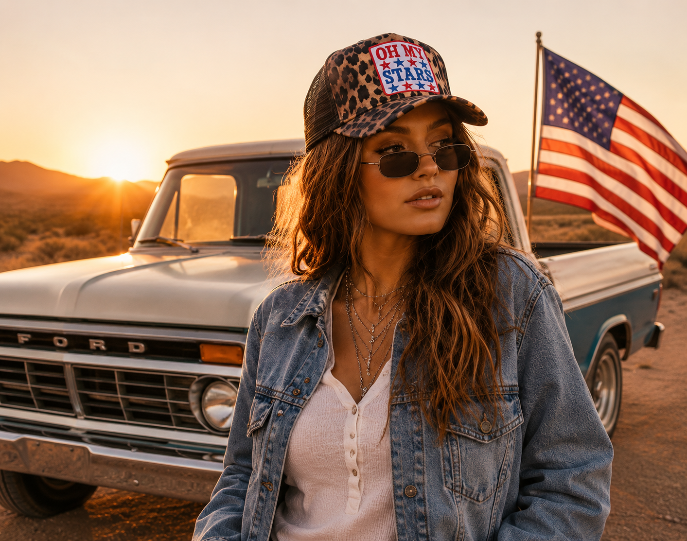 Woman wearing a cap with an American flag design, sunglasses, and denim jacket standing in front of a vintage Ford truck with an American flag in a desert setting.