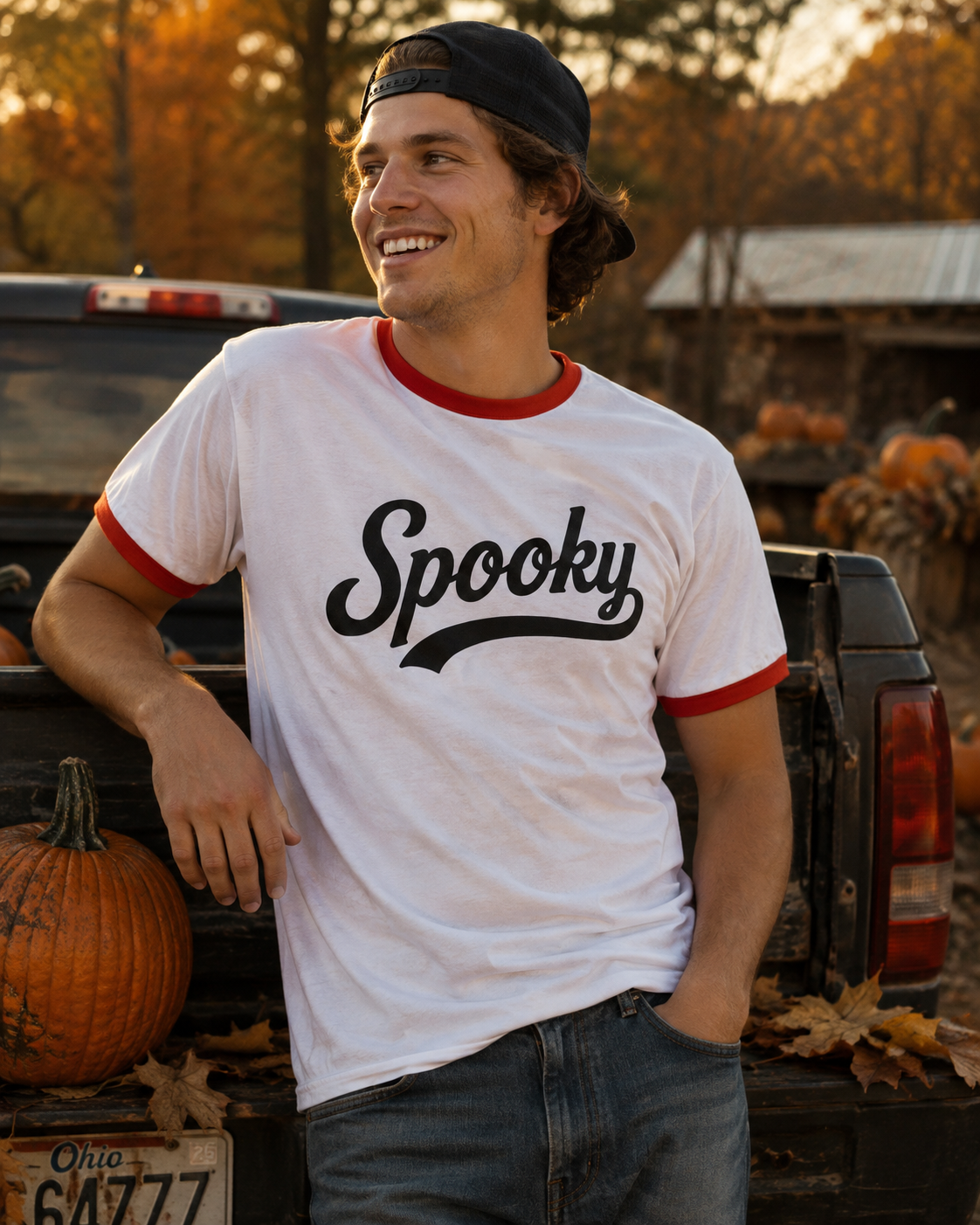 A man wearing a white ringer t-shirt with red trim and 'Spooky' printed in black script, leaning against the tailgate of a pickup truck with pumpkins in an autumn outdoor setting.