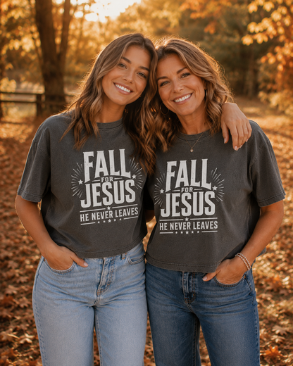 Two women standing outdoors in a wooded area with fallen leaves, wearing matching dark gray graphic T-shirts that read 'FALL FOR JESUS' and 'HE NEVER LEAVES'.
