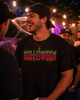 A man at a Halloween party wearing a black t-shirt with 'HALLOWEEN HALLOWEEN' printed in a colorful gradient font, paired with a black baseball cap and a silver cross necklace.