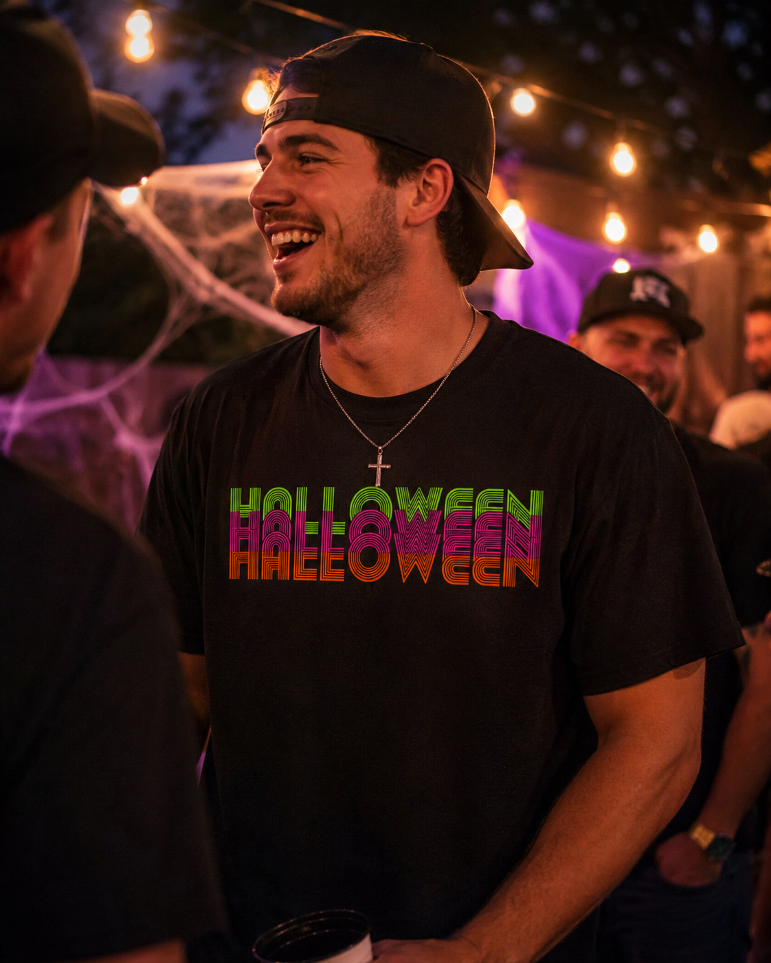 A man at a Halloween party wearing a black t-shirt with 'HALLOWEEN HALLOWEEN' printed in a colorful gradient font, paired with a black baseball cap and a silver cross necklace.