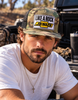 Man wearing a camo flat bill trucker hat with black mesh and a Like a Rock Jesus patch, seated beside a pickup truck.⠋