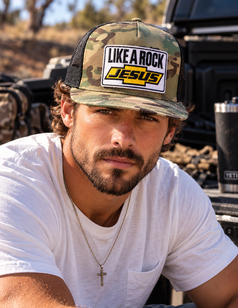 Man wearing a camo flat bill trucker hat with black mesh and a Like a Rock Jesus patch, seated beside a pickup truck.⠋