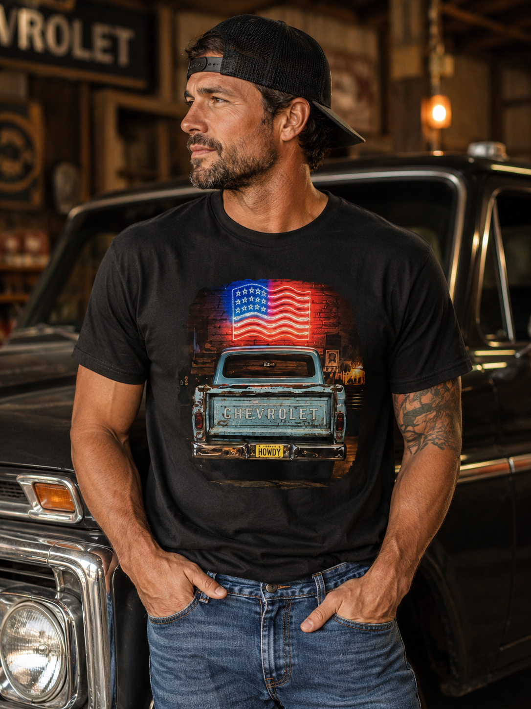 A man wearing a black graphic t-shirt with a vintage Chevrolet truck and American flag design, standing in front of a classic car in a garage setting.