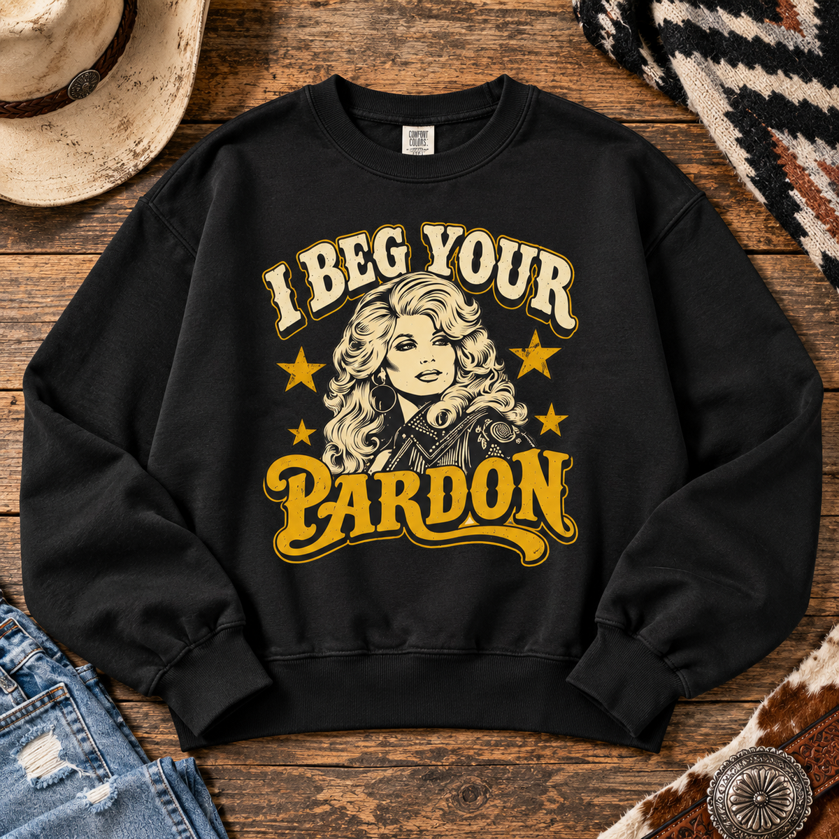 A black crewneck sweatshirt with a western-themed graphic reading 'I Beg Your Pardon' in cream and yellow lettering, featuring an illustration of a woman with blonde hair and stars. The sweatshirt is displayed in a flat-lay arrangement on a rustic wooden background with a cowboy hat, jeans, and a patterned textile.