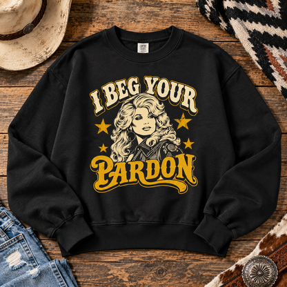 A black crewneck sweatshirt with a western-themed graphic reading 'I Beg Your Pardon' in cream and yellow lettering, featuring an illustration of a woman with blonde hair and stars. The sweatshirt is displayed in a flat-lay arrangement on a rustic wooden background with a cowboy hat, jeans, and a patterned textile.