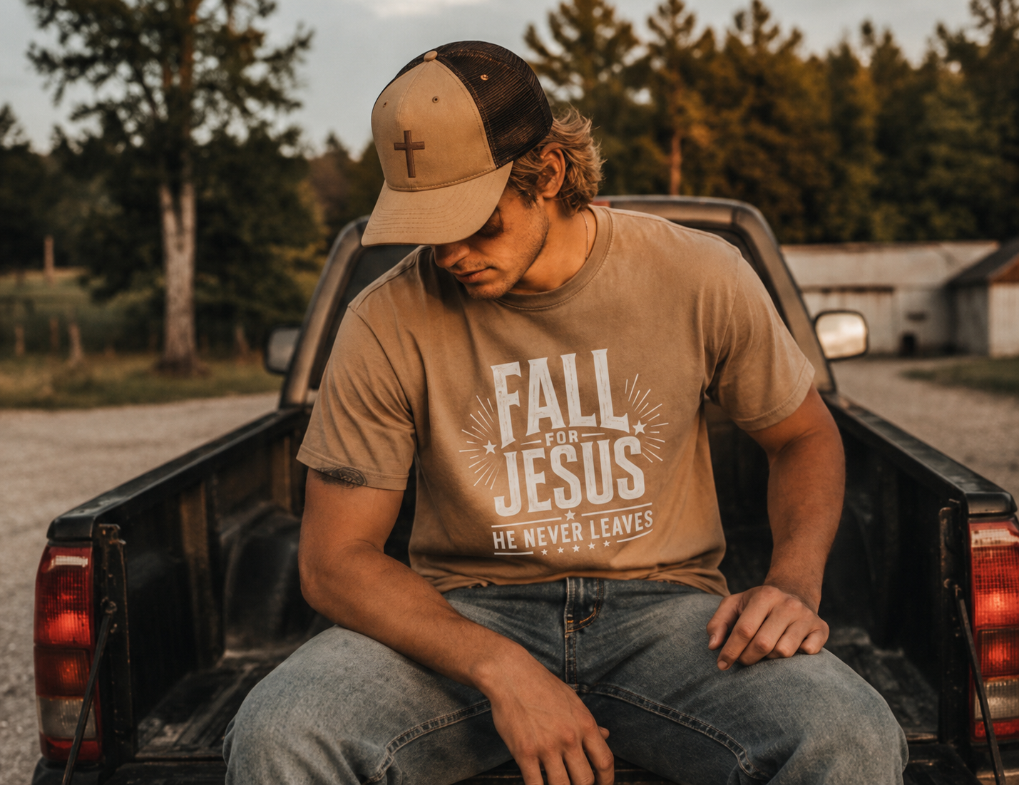 A man sitting in the bed of a pickup truck wearing a tan t-shirt with a white graphic that reads 'FALL FOR JESUS HE NEVER LEAVES' and a tan and black trucker hat with a cross on the front.