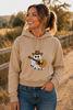 A woman wearing a beige hoodie with a cartoon ghost playing guitar wearing a cowboy hat, paired with blue jeans, standing on a dirt path outdoors.