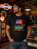 A man wearing a black graphic t-shirt with a vintage pickup truck and American flag design, standing in a retro-themed garage setting with neon signs and a classic car.