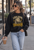 A woman walking on a city sidewalk wearing a black crewneck sweatshirt with a vintage-style graphic that reads 'I Beg Your Pardon' in yellow and cream, paired with light-wash blue jeans.