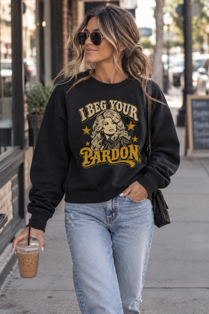 A woman walking on a city sidewalk wearing a black crewneck sweatshirt with a vintage-style graphic that reads 'I Beg Your Pardon' in yellow and cream, paired with light-wash blue jeans.