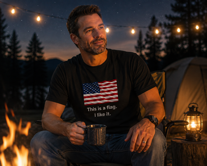 A man sitting by a campfire at night wearing a black t-shirt with a graphic of the American flag and the text 'This is a flag. I like it.
