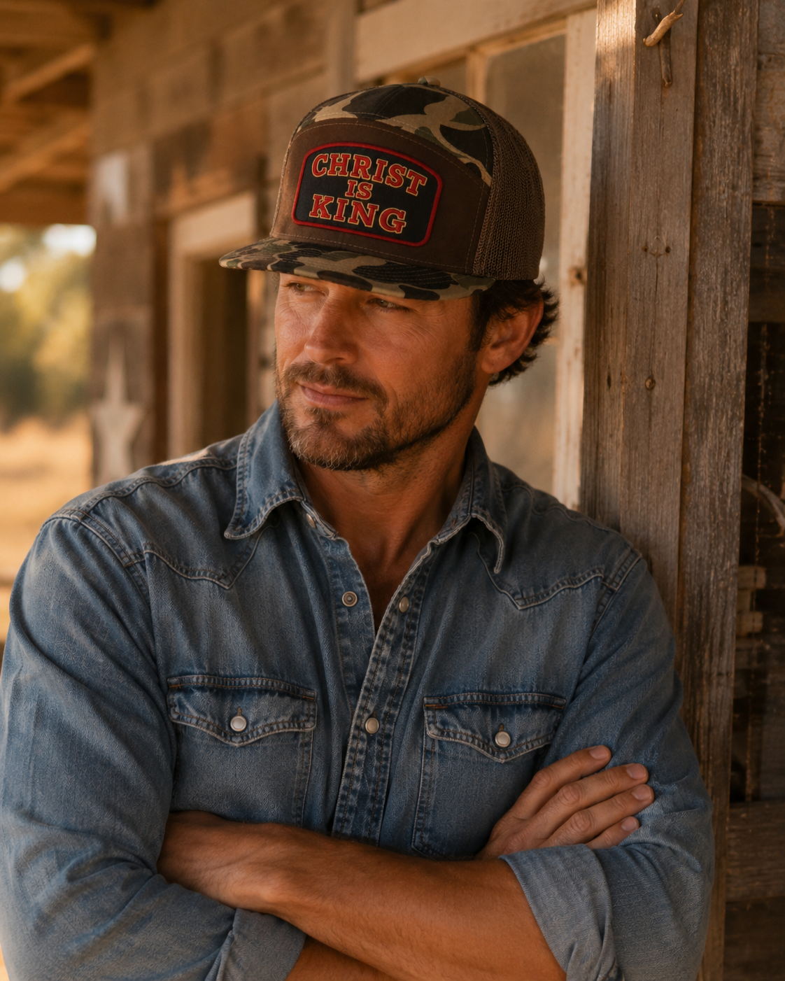A man wearing a camouflage trucker hat with 'CHRIST IS KING' embroidered on the front, paired with a blue denim shirt, leaning against a rustic wooden porch.