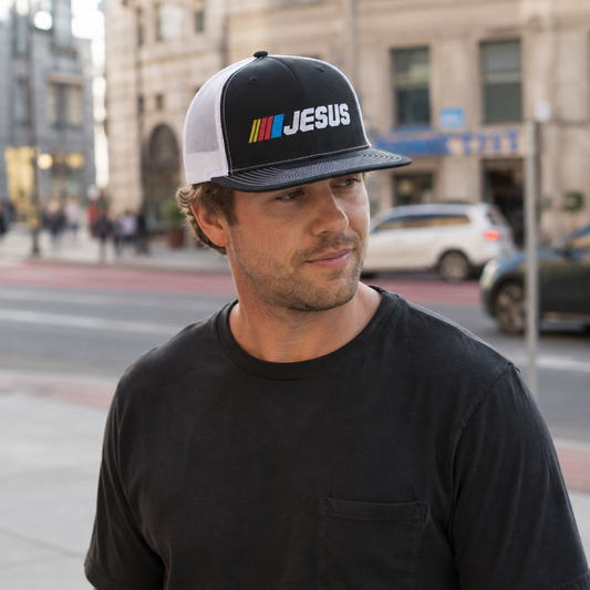 A man wearing a black and white trucker hat with a colorful 'JESUS' graphic and a black t-shirt, standing on a city street with buildings and cars blurred in the background.