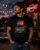 A man wearing a black t-shirt with a graphic of a vintage Chevrolet truck and an American flag, standing in front of a rustic Chevrolet sign at dusk.