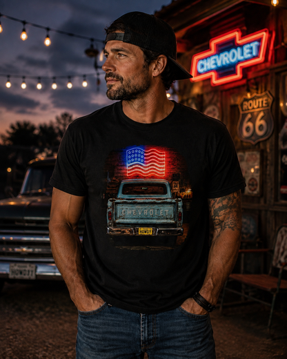 A man wearing a black t-shirt with a graphic of a vintage Chevrolet truck and an American flag, standing in front of a rustic Chevrolet sign at dusk.