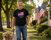 A man walking a golden retriever on a suburban sidewalk, wearing a black t-shirt with a graphic of the American flag and the text 'This is a flag I like it.'.