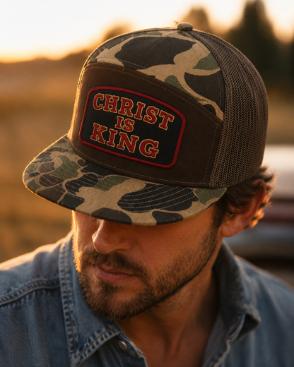 A man wearing a camouflage trucker hat with a front patch that reads 'CHRIST IS KING' in red letters, photographed outdoors at sunset.