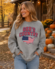 A woman wearing a heather gray crewneck sweatshirt with a 'God's Country' graphic featuring the American flag and a US map outline, set against an autumn outdoor background with pumpkins and a rustic cabin.