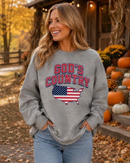 A woman wearing a heather gray crewneck sweatshirt with a 'God's Country' graphic featuring the American flag and a US map outline, set against an autumn outdoor background with pumpkins and a rustic cabin.