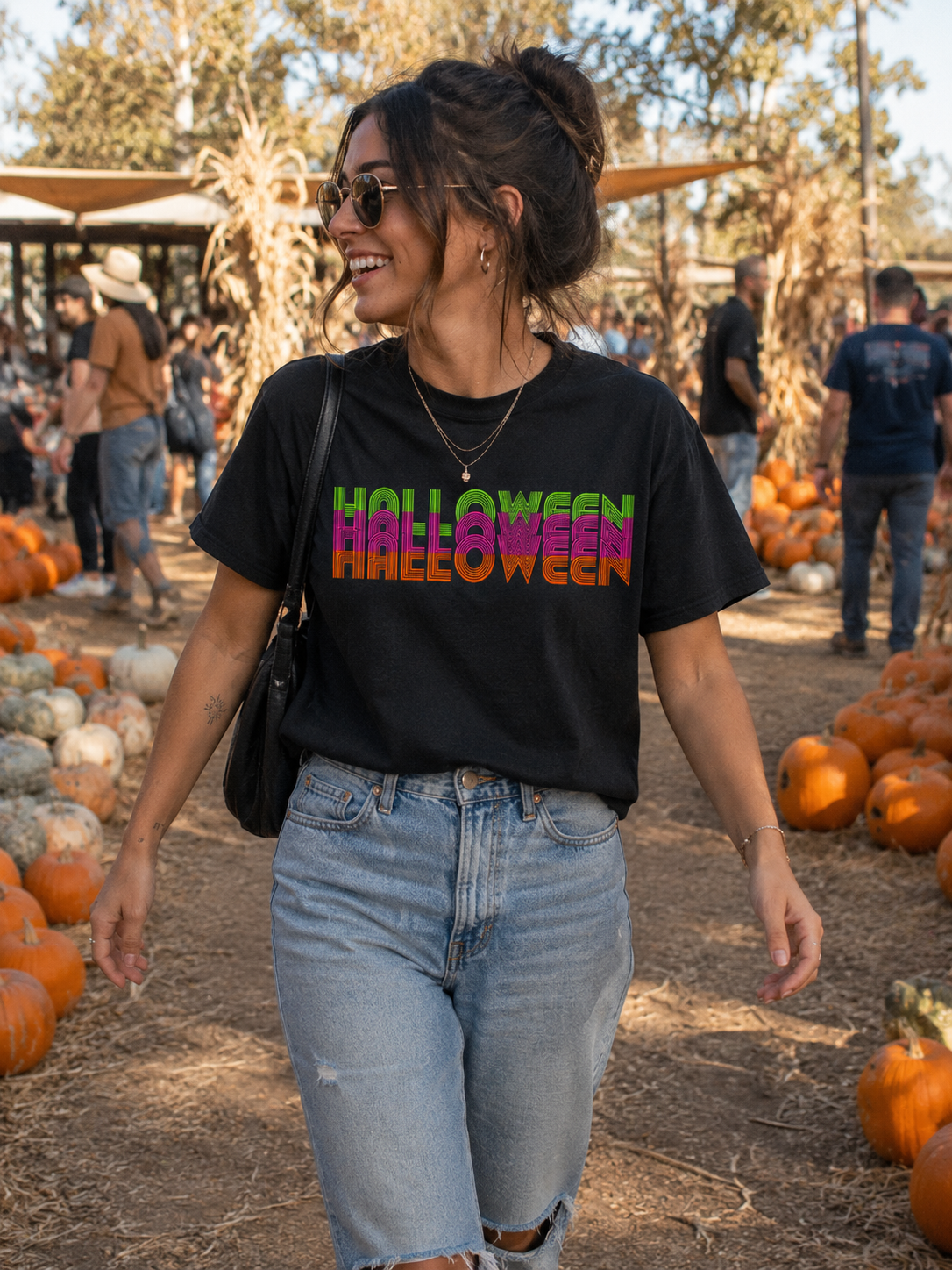 A woman walking in a pumpkin patch wearing a black graphic t-shirt with 'HOLLYWEEN HALLOWEEN' printed in a colorful gradient font, paired with light-wash blue jeans.